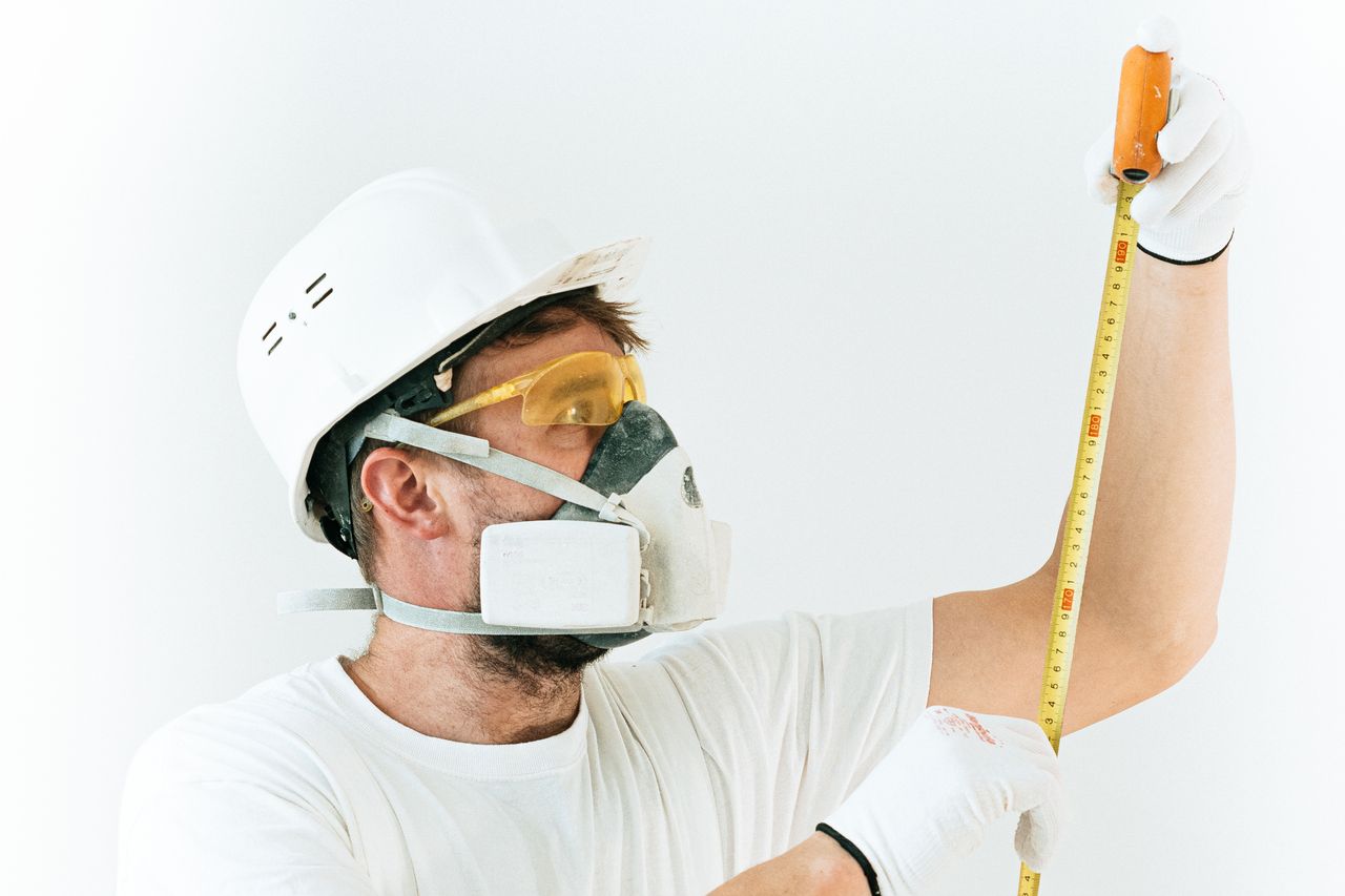 Construction worker using protective gear while measuring with a tape measure indoors.